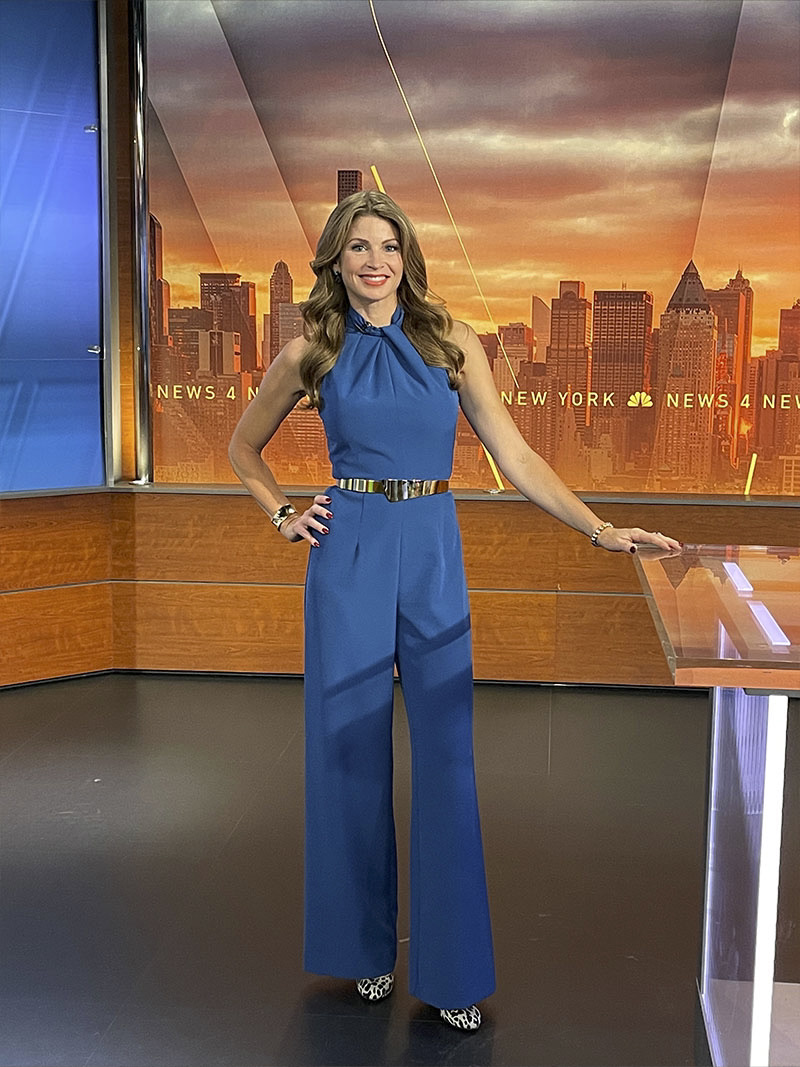 Jen on set, with one hand on her hip and the other on the anchor desk, smiling into camera wearing an elegant, dark teal jump suit.