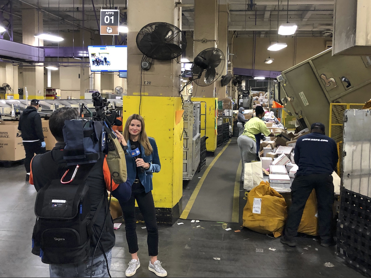 Jen anchoring in the field at a shipping warehouse, surrounded by workers and package bins, in jeans in a blue shaded velvet blazer.
