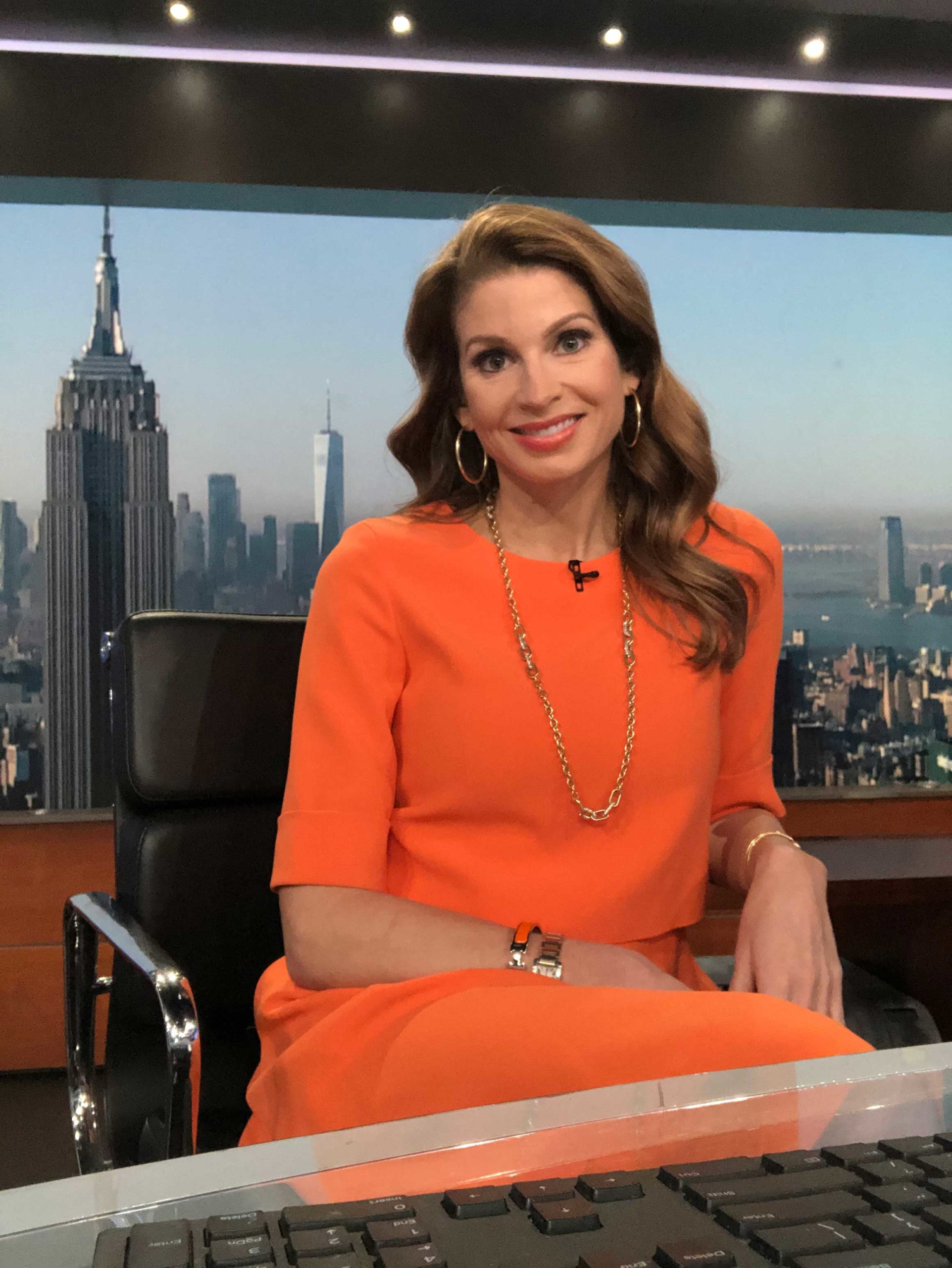Jen on set, sitting behind the anchor desk, smiling into camera in an orange dress.