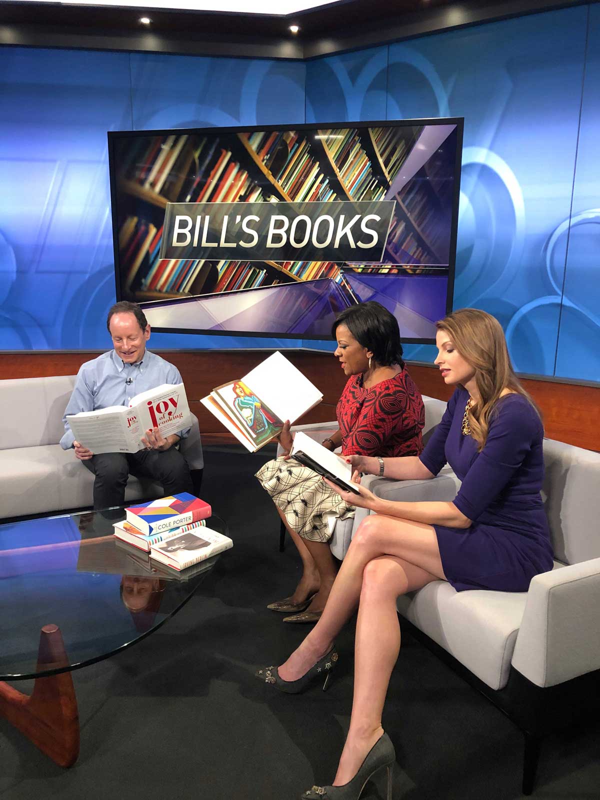 Jen on set with a colleague and a guest, sitting on white sofas reading. The video display behind reads "Bill's Books"